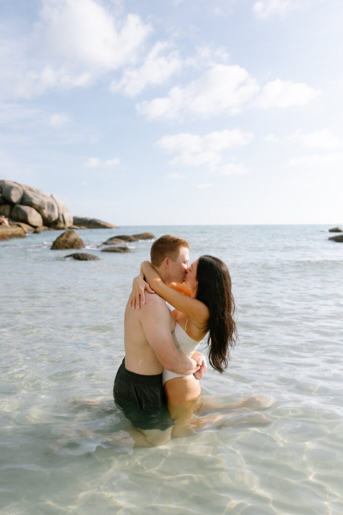 A couple embraces and kisses while kneeling in shallow, clear ocean water near large rocks under a partly cloudy sky.