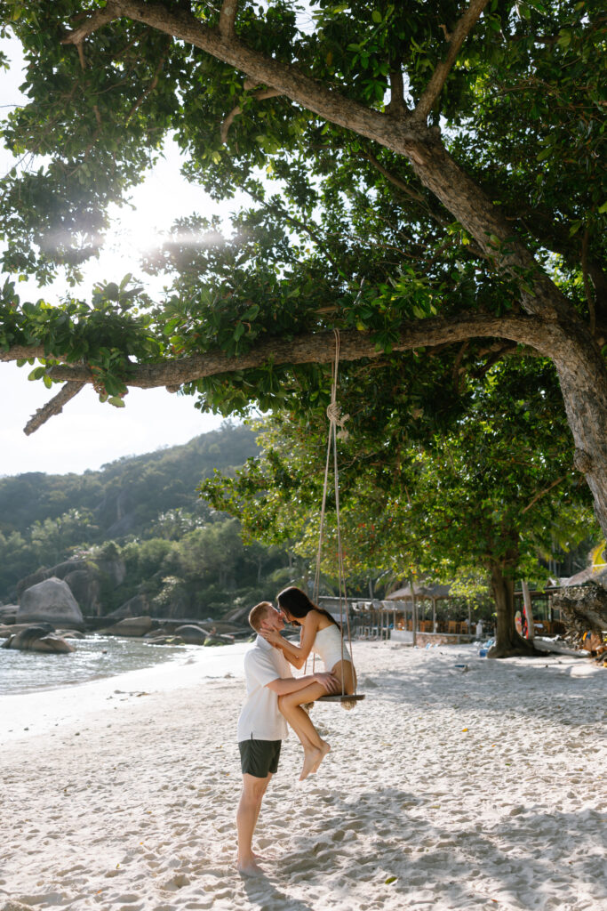 A couple enjoys a moment on a swing attached to a large tree on a sandy beach, surrounded by lush greenery and hills in the background, with sunlight filtering through the leaves.