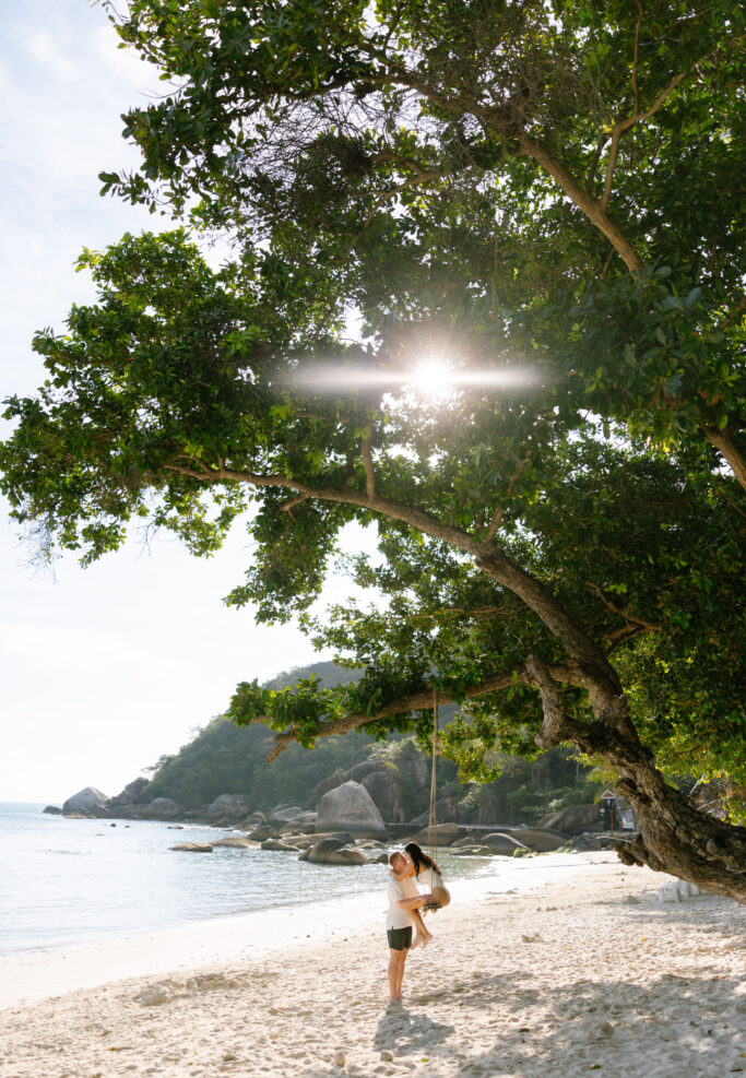 A woman swings on a rope swing hanging from a large tree on a sandy beach, with sunlight shining through the leaves and the sea and rocks in the background.