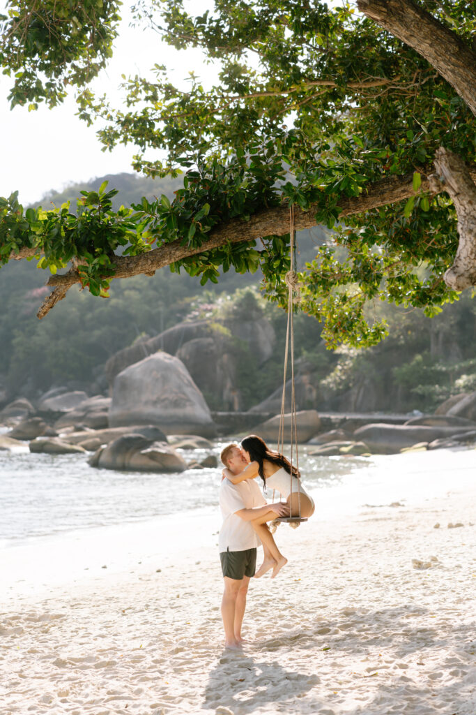 A man on a sandy beach gently pushes a child sitting on a rope swing hanging from a tree branch. The background has large rocks, greenery, and distant hills by the water.