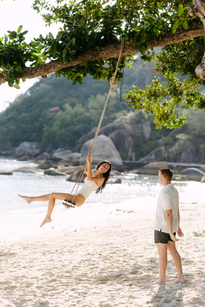 A woman in a swimsuit happily swings on a rope swing attached to a tree branch on a sandy beach, while a man in a white shirt and shorts stands nearby, watching her. Rocky hills and water are visible in the background.