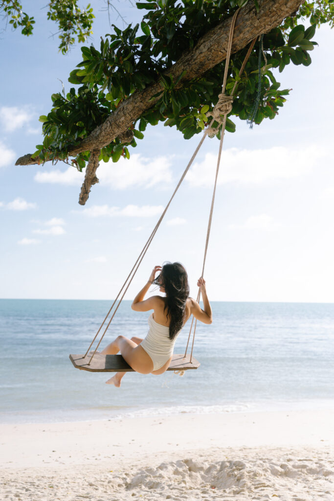 A woman in a white swimsuit on a swing on a beach.