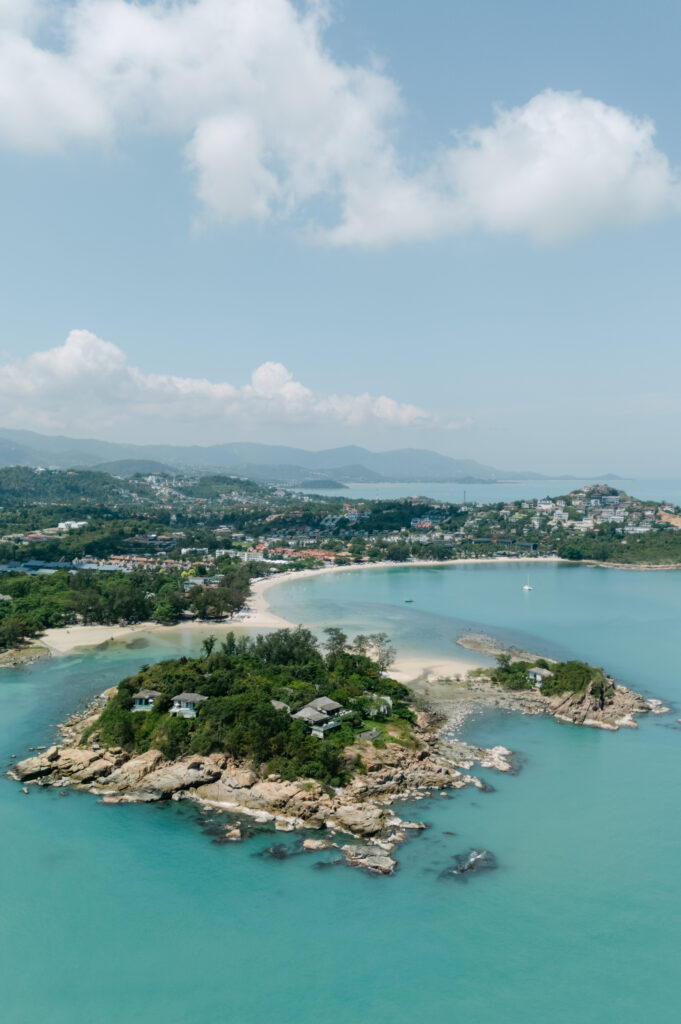 Aerial view of a small rocky island with lush greenery, turquoise water, sandy beaches, and a coastal town with hills in the background under a partly cloudy sky.