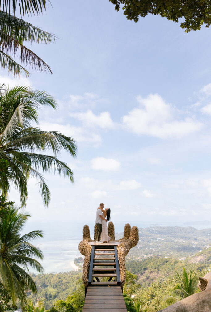 A couple embraces at the end of a wooden walkway shaped like giant hands, overlooking a lush landscape, coastline, and blue sky with scattered clouds. Palm trees frame the scene.