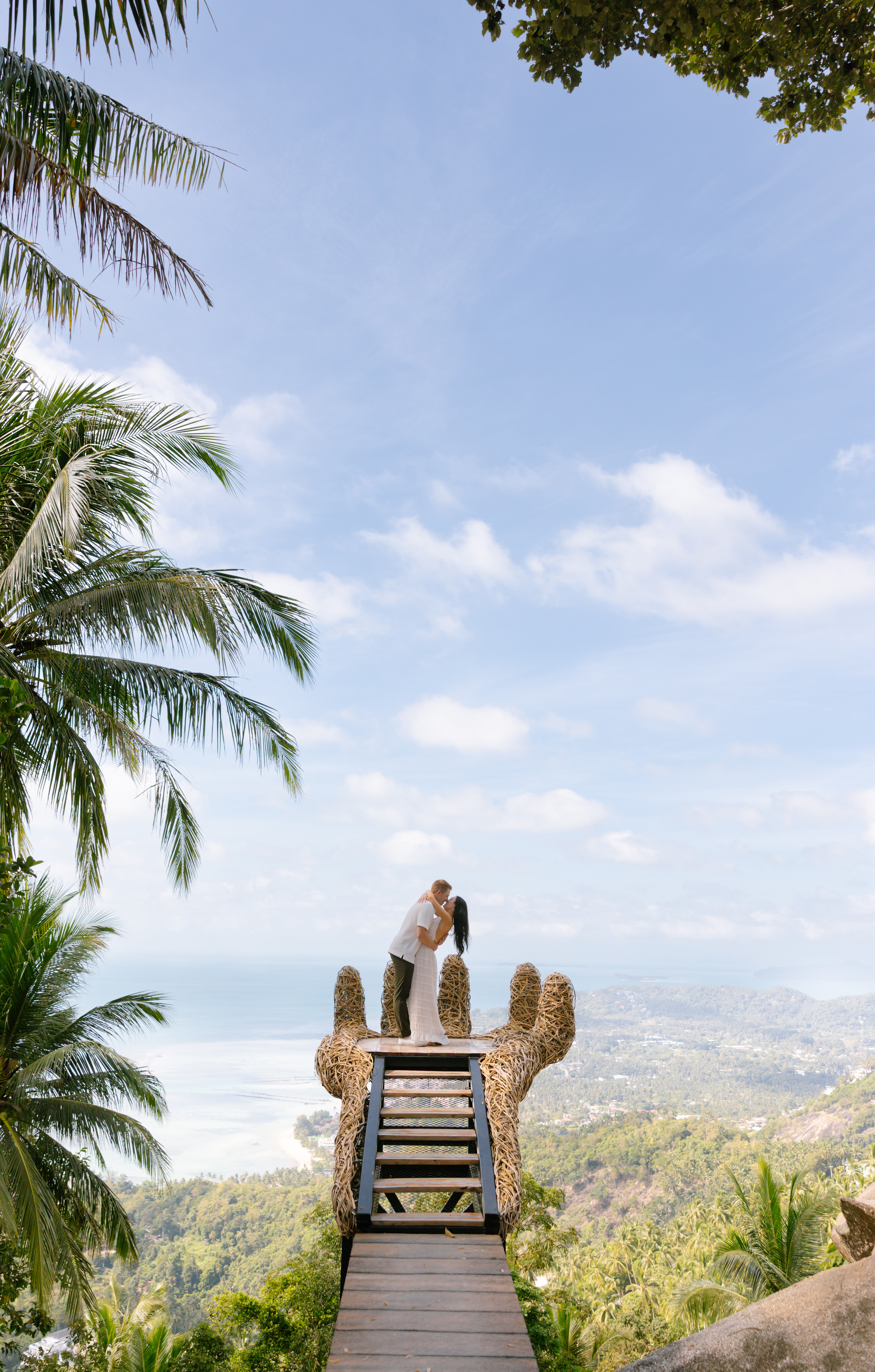 A couple stands on a wooden platform shaped like a giant hand, surrounded by tropical trees, overlooking the ocean and a lush landscape under a bright blue sky.