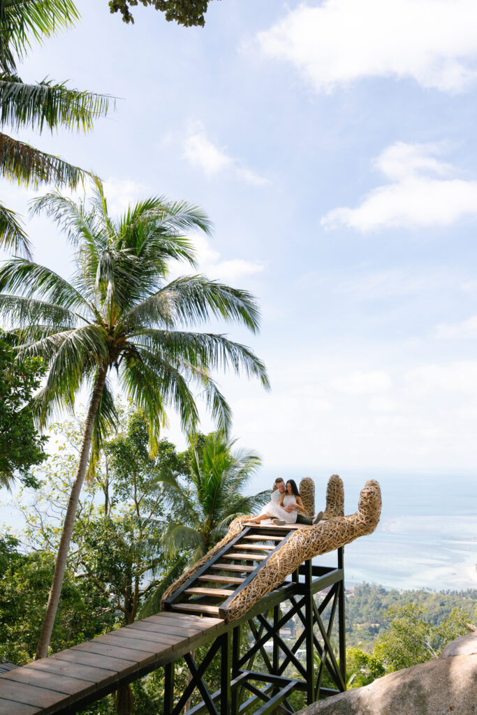 A couple sits together on a large hand-shaped platform overlooking lush tropical trees, with a scenic ocean view in the background under a bright, partly cloudy sky.
