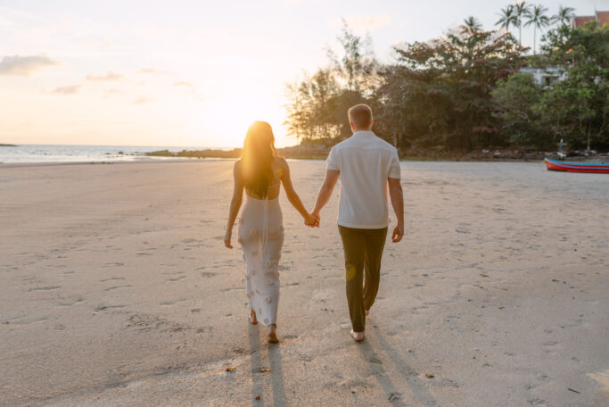 A couple holding hands walks barefoot on a sandy beach at sunset, with the sun low on the horizon and trees in the background.