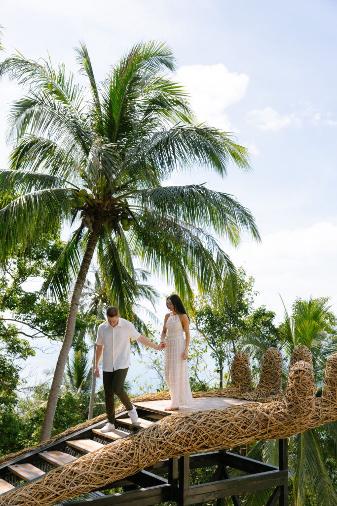 A man and woman hold hands while walking down a woven, nest-shaped structure with railings, surrounded by lush green palm trees and tropical foliage under a bright sky.