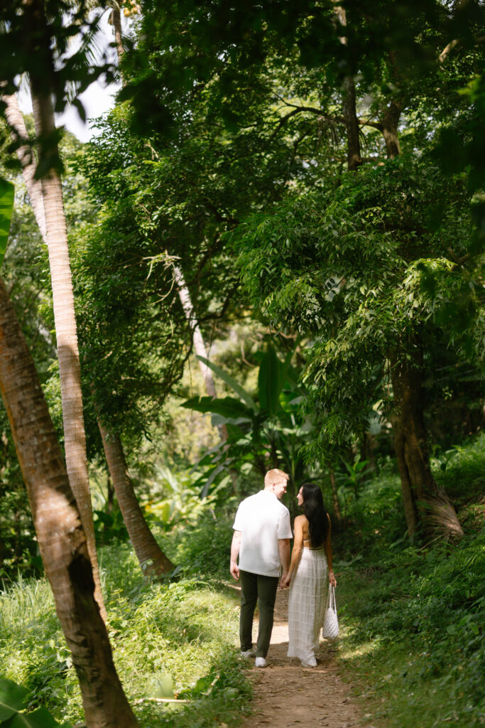 A couple walks hand-in-hand along a narrow, sunlit path surrounded by lush green trees and vegetation in a tropical forest.
