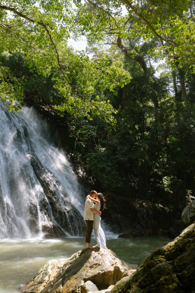 A couple embraces on a rock in front of a waterfall, surrounded by lush green trees and sunlight filtering through the leaves.