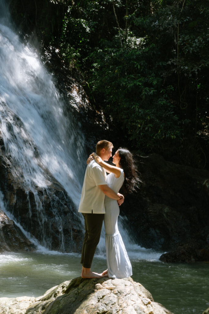 A couple stands barefoot on a rock in front of a waterfall, embracing and gazing into each others eyes, surrounded by lush greenery and sunlight.