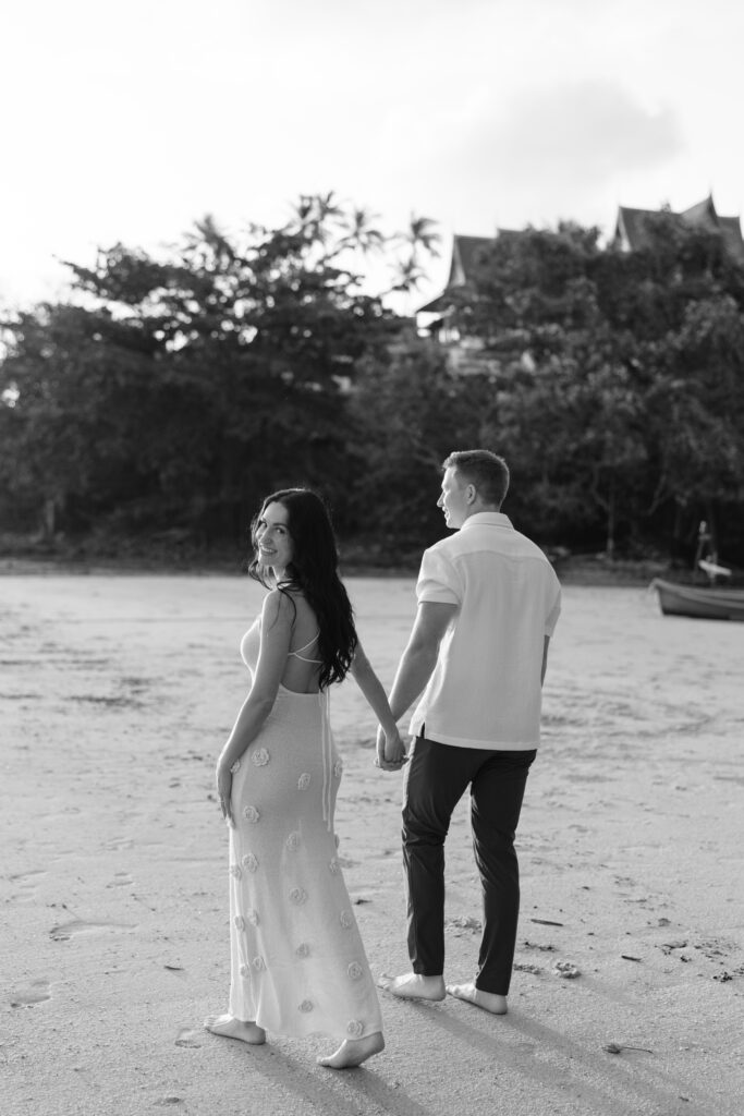 A couple walks hand in hand on a sandy beach, looking back and smiling. Trees and buildings are visible in the background. The photo is in black and white.