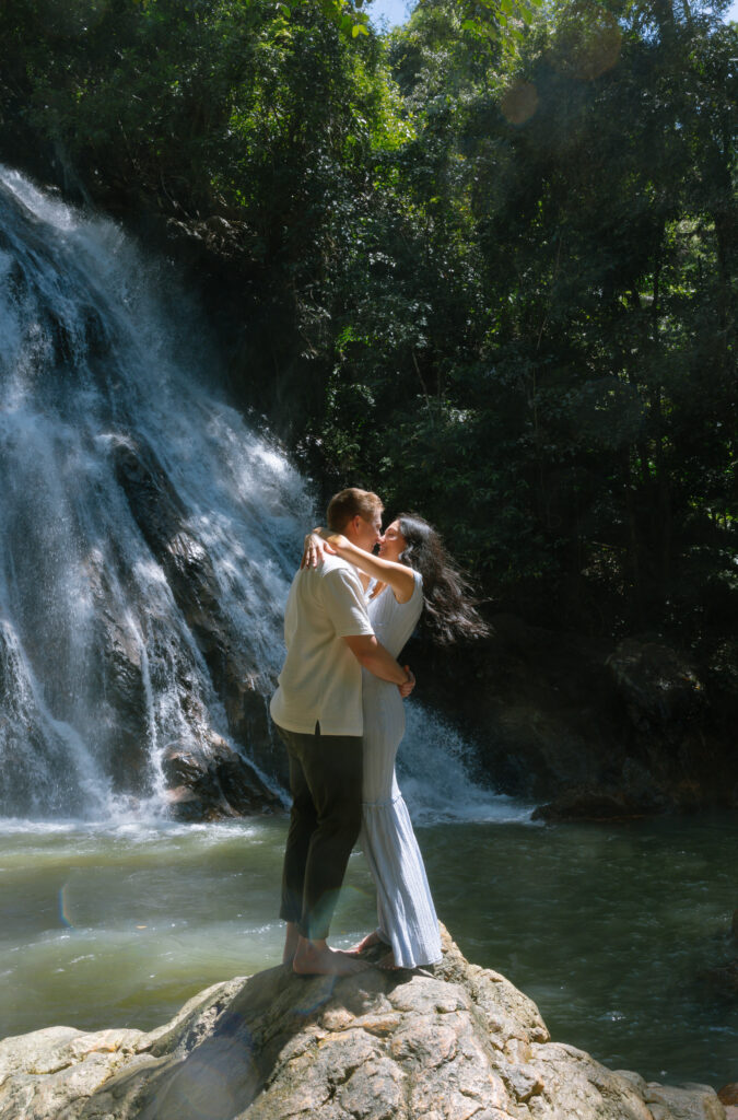 A couple embraces and kisses on a large rock in front of a waterfall, surrounded by lush green forest and sunlight filtering through the trees.
