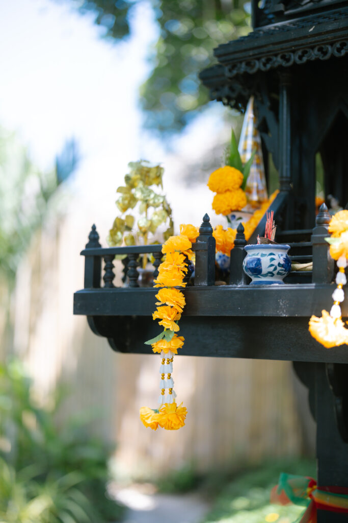 A close-up of a black spirit house decorated with yellow marigold flower garlands, a blue-and-white ceramic incense holder, and other offerings, set outdoors with greenery and a bamboo fence in the background.
