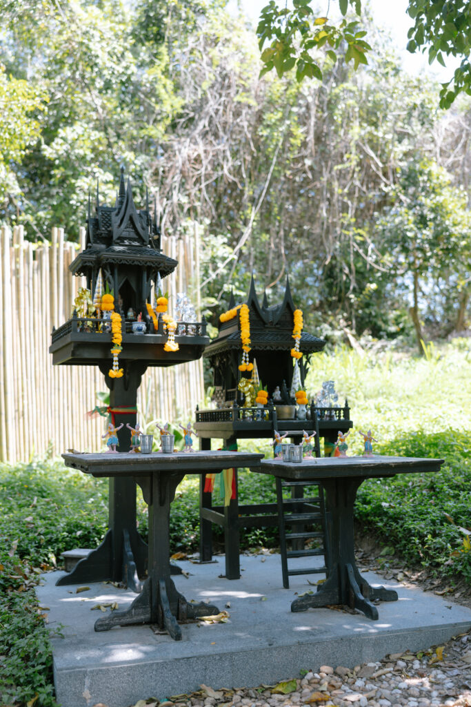 A Thai spirit house shrine with intricate black structures decorated with yellow marigold garlands, small figurines, and offerings, set outdoors amid greenery and sunlight.