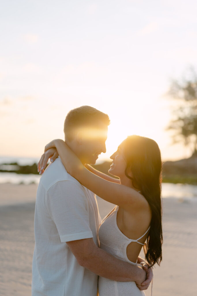 A couple embraces on a sandy beach at sunset, smiling at each other with warm sunlight glowing behind them. The woman has long dark hair and wears a sleeveless dress; the man wears a short-sleeved shirt.