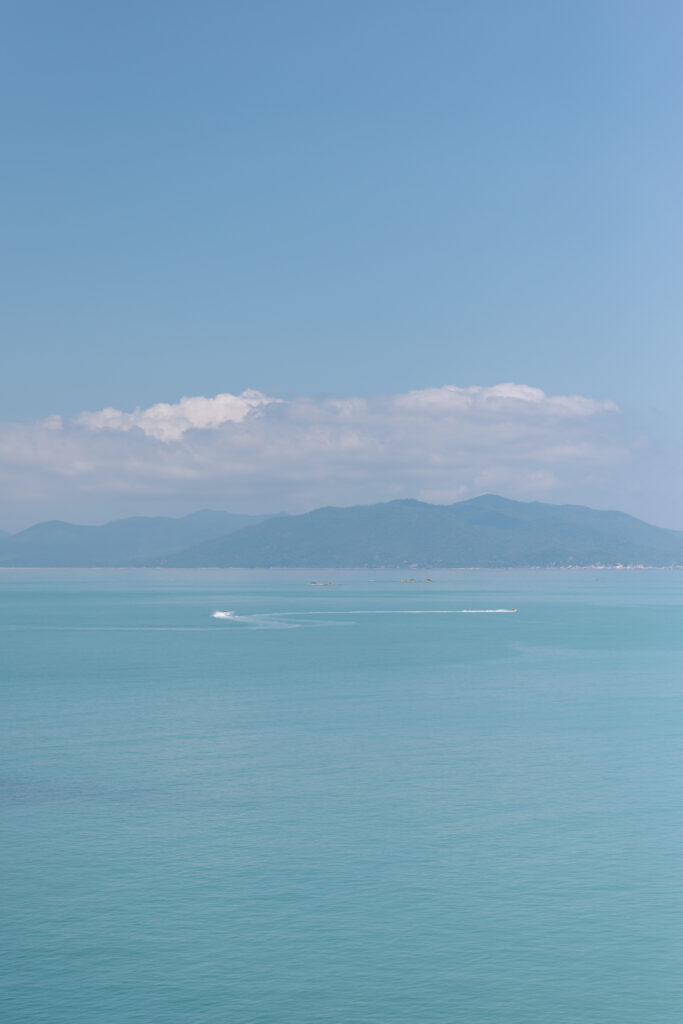 Calm turquoise sea under a clear blue sky with a few scattered clouds. Distant mountains stretch across the horizon, and faint white waves appear on the waters surface.