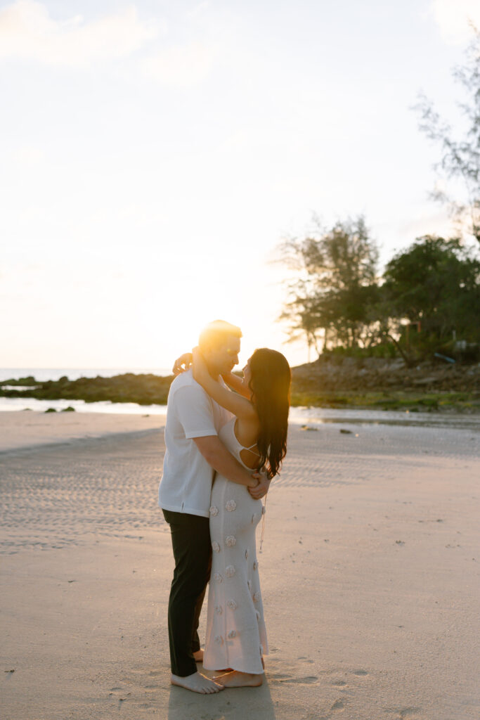 A couple embraces on a sandy beach at sunset, with sunlight softly glowing behind them. Trees and rocks line the shore in the background, creating a peaceful, romantic scene.