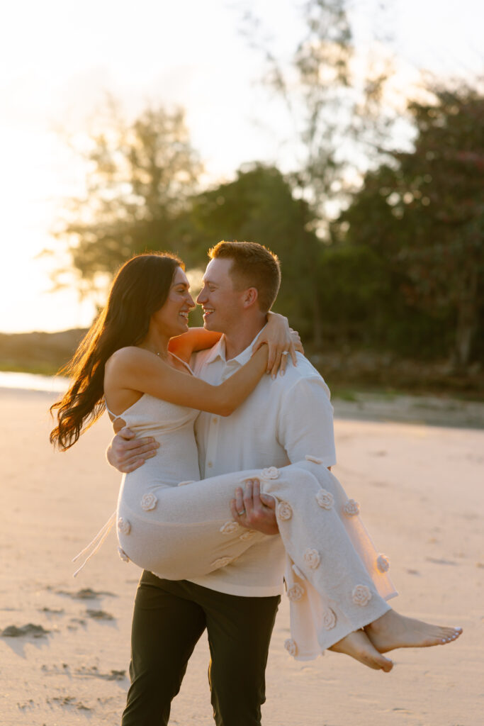A man in a white shirt holds a smiling woman in a white dress in his arms on a sandy beach at sunset, with trees in the background.