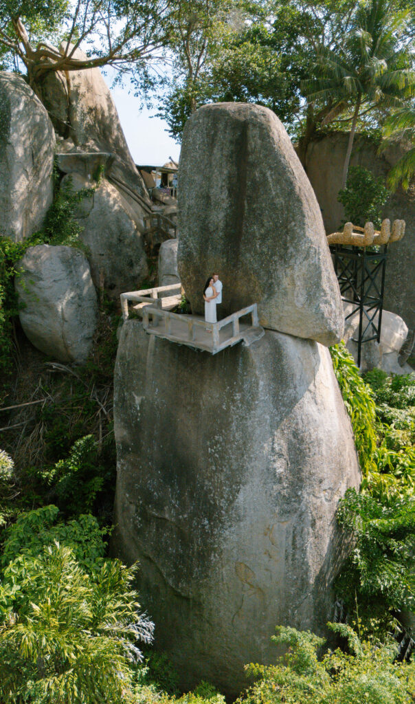 A person in white clothing stands on a small, fenced platform built atop a tall, narrow rock surrounded by lush greenery and large boulders.