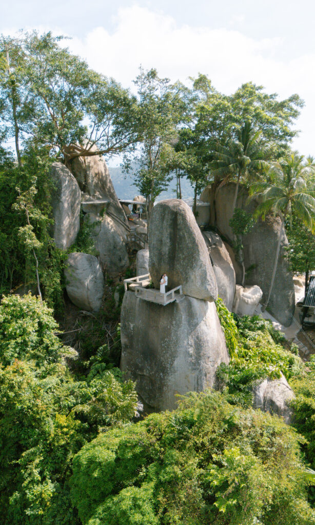 A person stands on a small white viewing platform built atop large, smooth boulders surrounded by green trees and dense foliage under a partly cloudy sky.