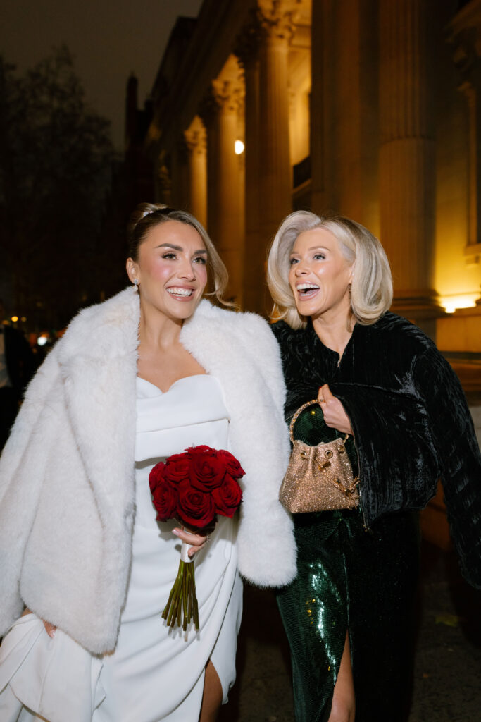 Two women walking outside at night, smiling. One wears a white dress and coat, holding red roses; the other wears a dark, sparkly outfit and carries a gold purse. Tall columns and warm lights are visible in the background.