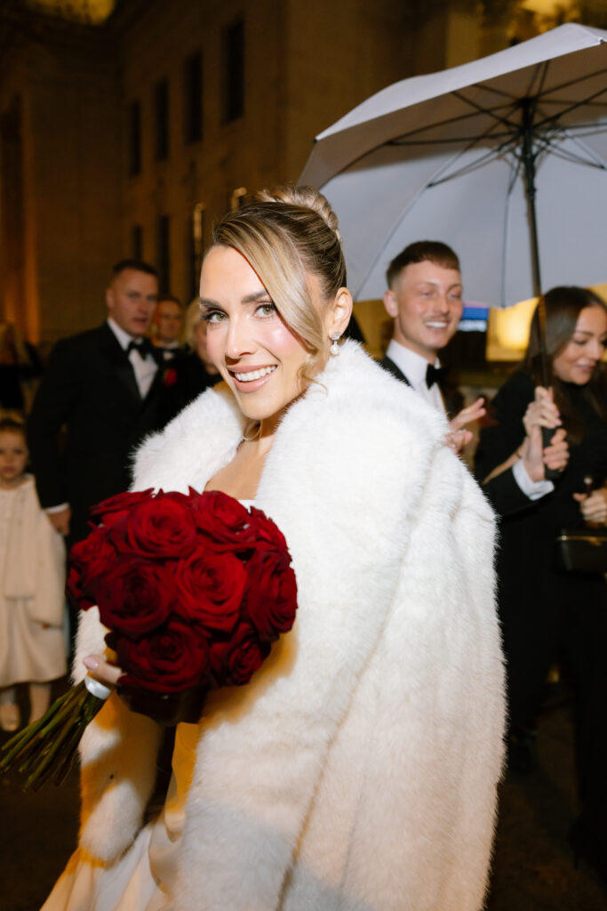 A bride in a white fur coat holds a bouquet of red roses and smiles at the camera. People in formal attire, including someone holding an umbrella, stand around her on a nighttime street.