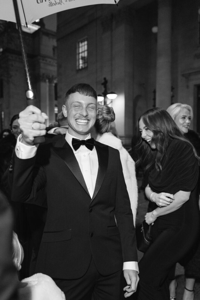 A man in a tuxedo smiles broadly and holds an umbrella at a nighttime outdoor event, surrounded by people dressed in formal attire. The scene is lively and joyful.