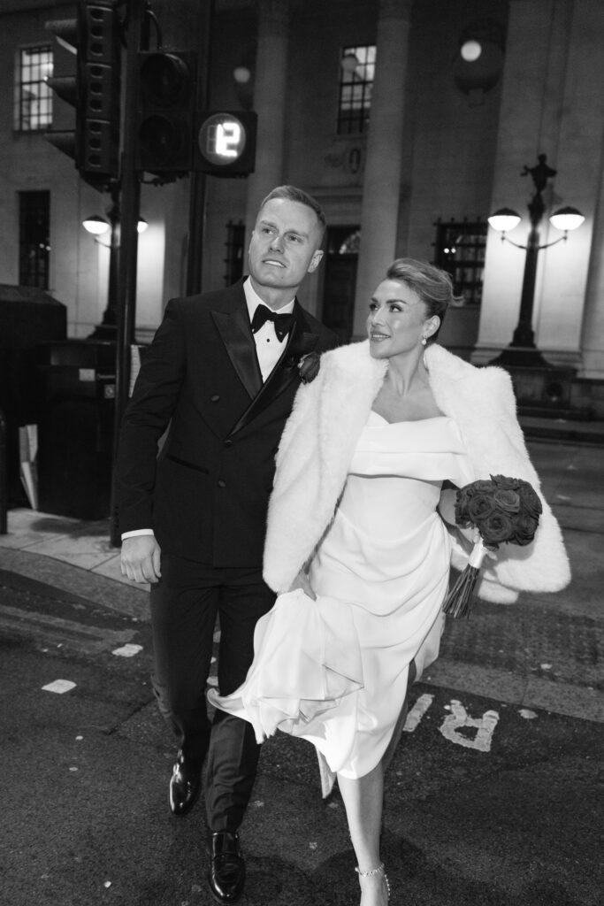 A couple dressed formally, with the woman in a white dress and fur coat holding a bouquet. The man wears a tuxedo. They walk together at night on a city street, appearing joyful and elegant.