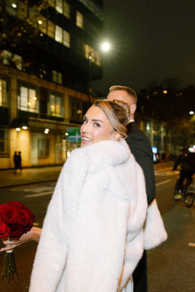 A woman in a white fur coat holding a bouquet of red roses looks back and smiles at the camera while standing on a city street at night with a man beside her.