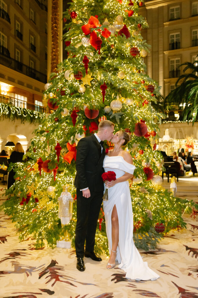 A bride and groom kiss in front of a tall, decorated Christmas tree with red ornaments and lights. The bride holds red roses and wears a white dress with a slit; the groom wears a black suit. The elegant room is festively lit.