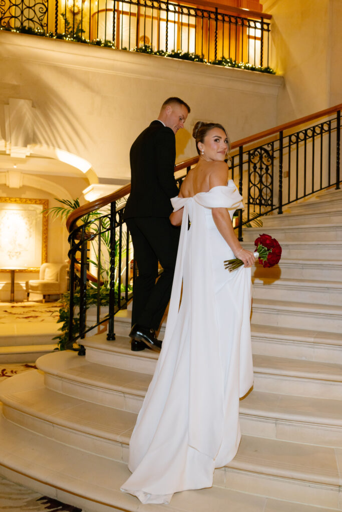 A bride in an off-shoulder white gown holds a red bouquet and looks back while standing on a grand staircase with a groom in a black suit, in an elegant indoor setting with warm lighting.