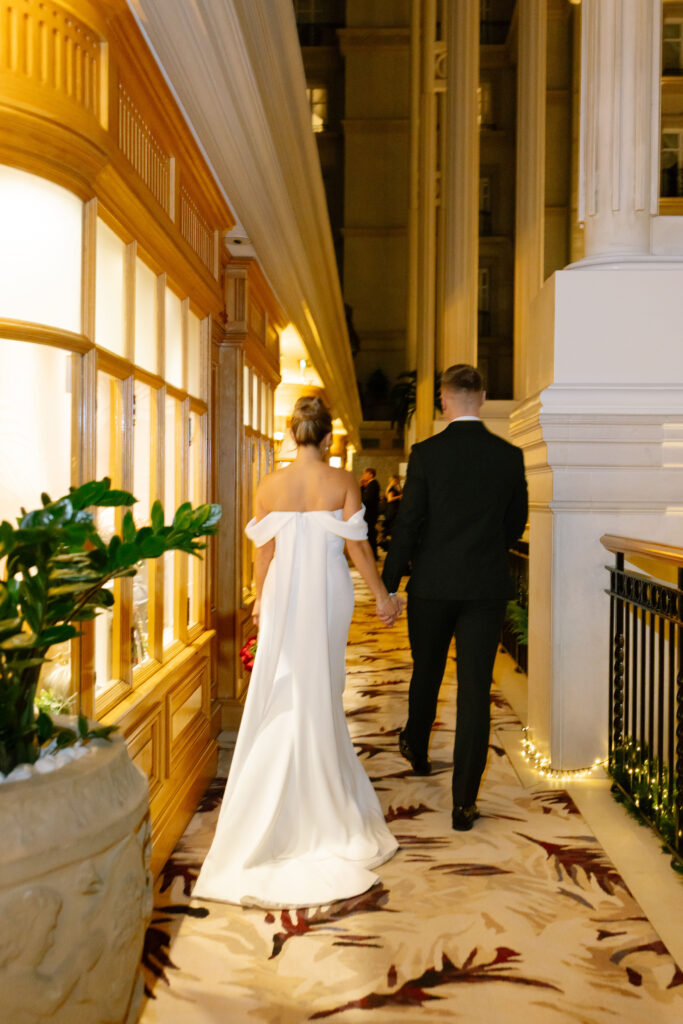 A bride in an off-the-shoulder white gown and a groom in a black suit walk hand-in-hand down a warmly lit hallway with patterned carpet and large decorative plants.