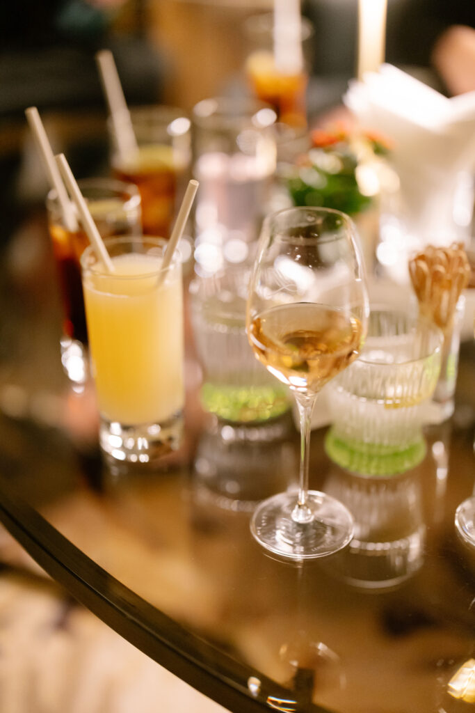 A close-up of a glass table with assorted drinks, including a glass of wine, a yellow cocktail, and other beverages with straws, surrounded by glasses and a blurred floral centerpiece in the background.