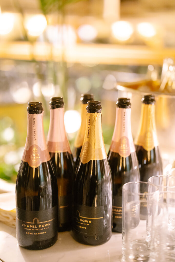 Six bottles of Chapel Down Rosé Reserve sparkling wine are arranged on a table, with empty glasses nearby. The background is softly blurred with warm lights, creating an elegant atmosphere.