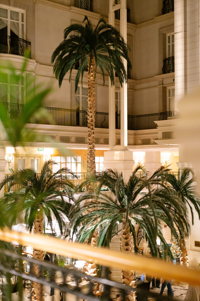 Tall artificial palm trees stand in the center of an elegant, well-lit indoor atrium with balconies and large windows on multiple floors. A gold railing is visible in the foreground.