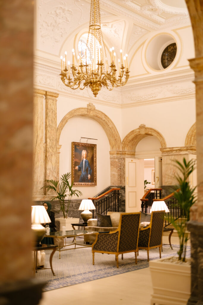 A luxurious hotel lobby with elegant furniture, a large gold chandelier, marble columns, indoor plants, and a portrait painting on the wall above a staircase. Warm lighting creates a welcoming atmosphere.