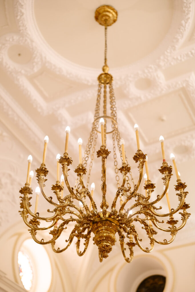 An ornate gold chandelier with multiple lit candles hangs from an elegant, decorative white ceiling with intricate molding details.