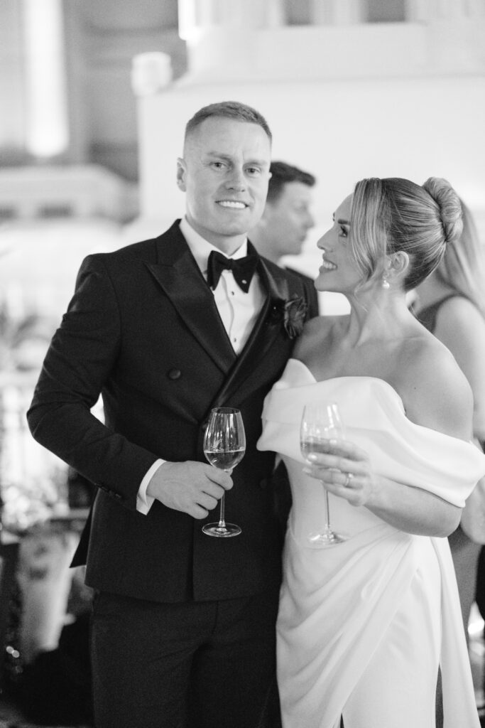 A man in a tuxedo and a woman in an off-the-shoulder white dress stand together at a formal event, smiling and holding wine glasses. The background is softly blurred with other guests visible.