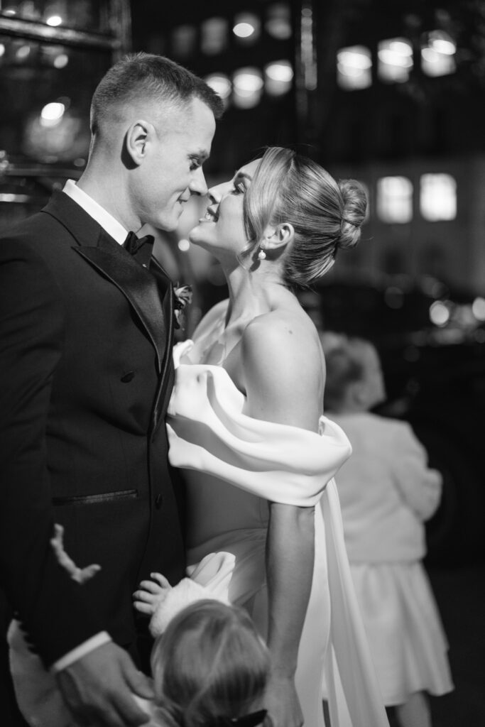 A bride and groom dressed formally share a joyful, intimate moment, smiling closely at each other. The scene is in black and white, with blurred people and lights in the background.