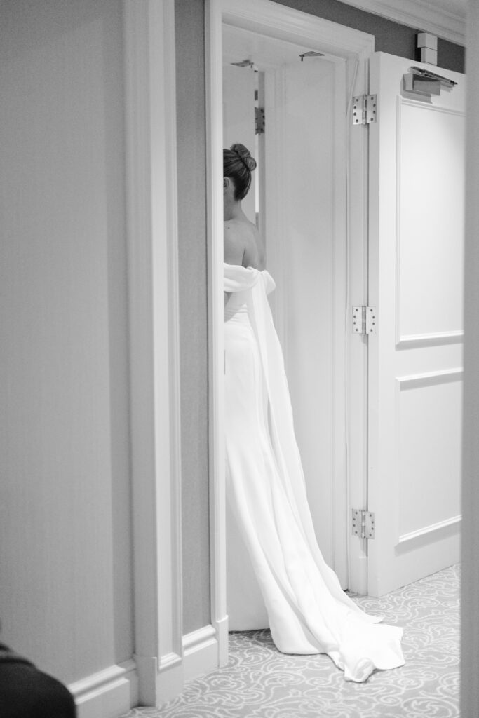 A bride in an off-the-shoulder white gown stands partially hidden behind a door, with her back turned and a long train trailing behind her in a softly lit hallway.
