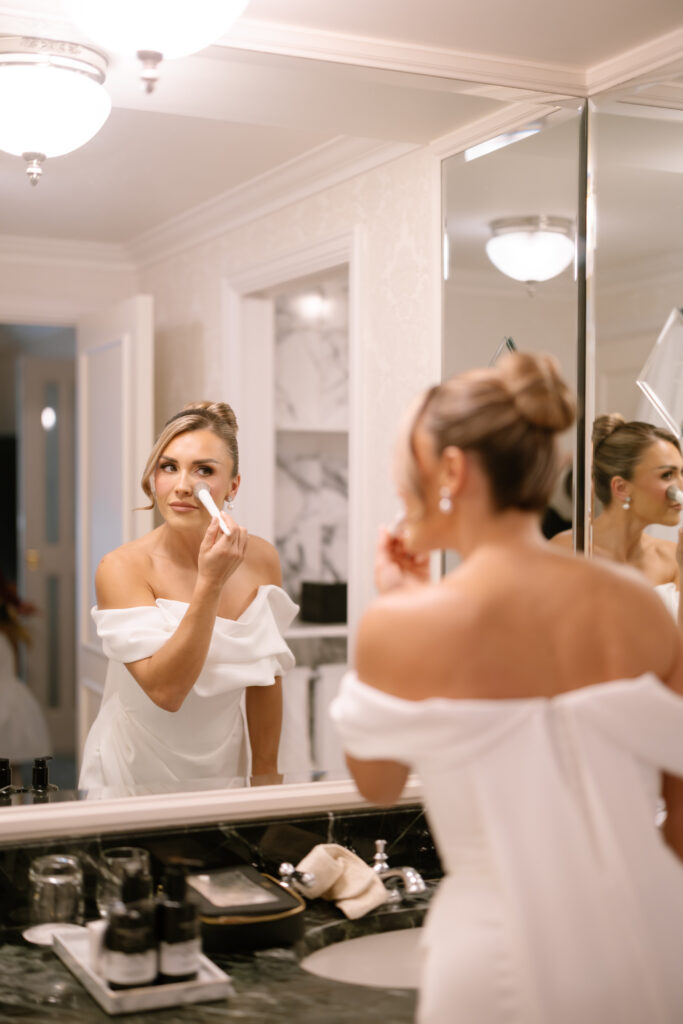 A woman in an off-the-shoulder white dress applies makeup with a brush while standing at a bathroom counter, looking into a large mirror. The bathroom is elegant and well-lit.
