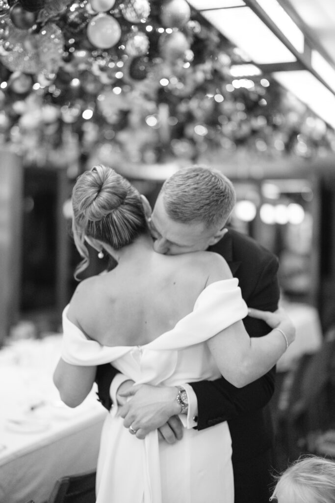 A black and white photo of a couple embracing closely; the man gently kisses the womans neck as she holds him, with festive decorations hanging above them in the background.