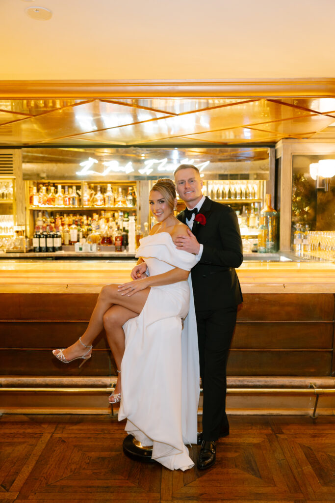 A bride in a white off-shoulder gown with a thigh-high slit sits on a bar stool, posing with a groom in a black tuxedo in front of a well-lit bar filled with bottles. Both are smiling and dressed for a formal occasion.