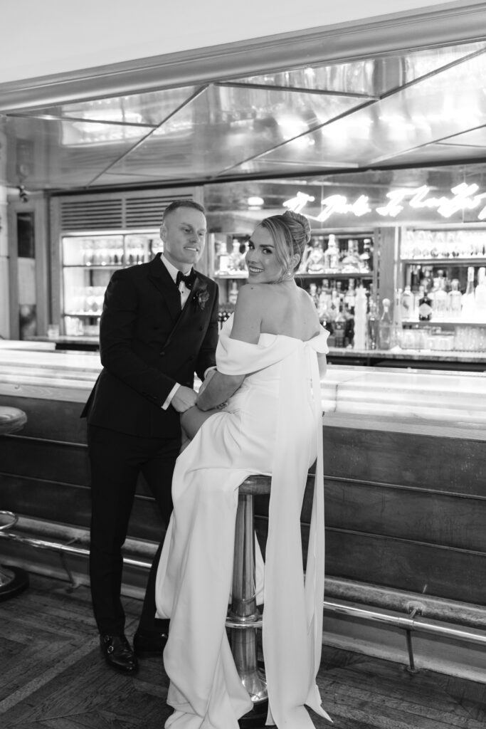 A bride in an off-the-shoulder gown and a groom in a tuxedo pose together at a bar, smiling and looking at the camera in an elegant, classic setting.