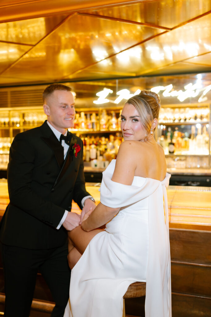 A bride in an off-the-shoulder white gown sits at a bar, smiling slightly at the camera, while a groom in a black tuxedo with a red boutonniere stands beside her. The background features warm lighting and shelves of bottles.