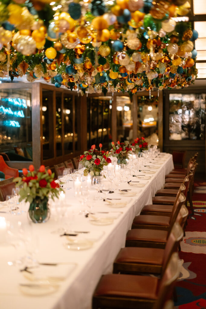 A long, elegant dining table set with white linens, wine glasses, candles, and red floral centerpieces, with colorful holiday ornaments and greenery hanging overhead in a warmly lit room.