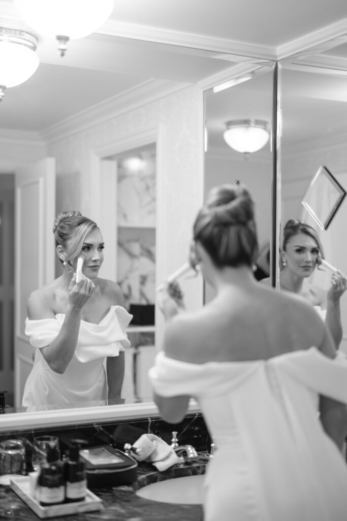 A woman in an off-shoulder dress stands at a bathroom mirror, applying makeup. Her hair is styled in an elegant updo, and the setting appears to be sophisticated and well-lit. The image is in black and white.