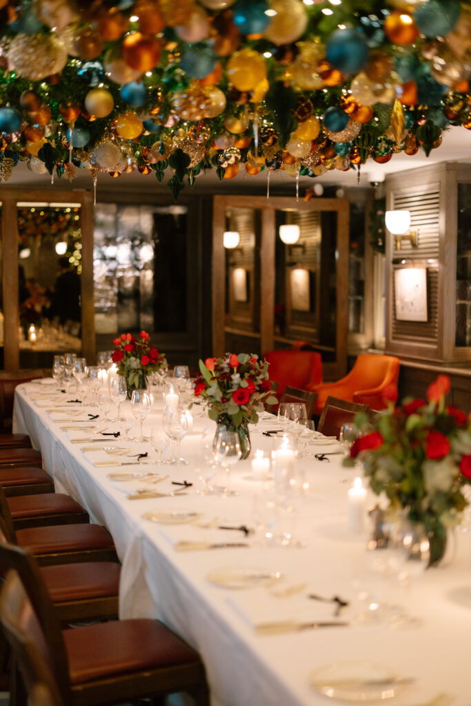 A long, elegant dining table with a white tablecloth is set for a formal event, decorated with candles and bouquets of red roses. Colorful ornaments and greenery hang densely from the ceiling above.