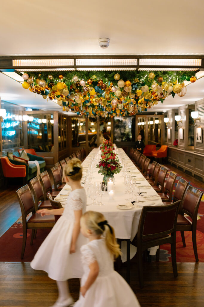 A long dining table set for an elegant event is decorated with flowers and surrounded by chairs. Ornate holiday ornaments hang from the ceiling. Two young girls in white dresses move near the table.